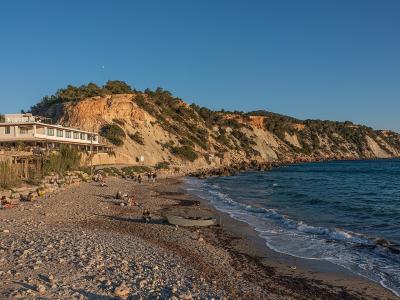A coastal scene showing a pebble beach, a white multi-story building built into a cliffside, and the sea under a clear blue sky.