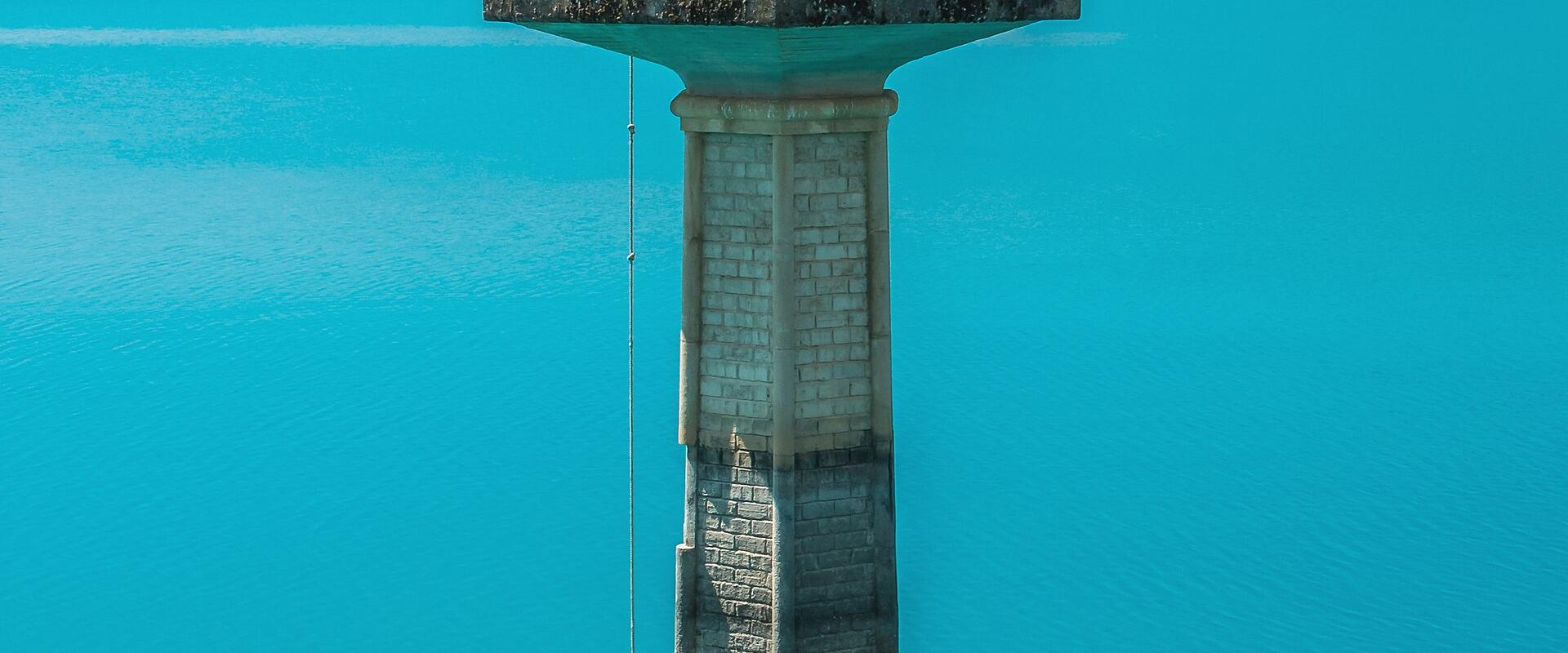 A stone watchtower or intake tower with a domed top and circular windows stands in the middle of a bright turquoise reservoir, with hills and mountains in the background.