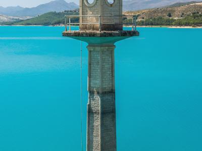 A stone watchtower or intake tower with a domed top and circular windows stands in the middle of a bright turquoise reservoir, with hills and mountains in the background.