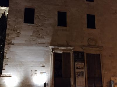 A night-time photo of a stone building facade with six dark rectangular windows, an entrance doorway, and a vertical banner sign next to the door.