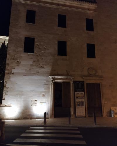 A night-time photo of a stone building facade with six dark rectangular windows, an entrance doorway, and a vertical banner sign next to the door.