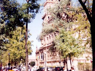 A street scene in Seville showing the Plaza de América with the distinctive Moorish Revival tower of the Museum of Popular Arts and Customs (Museo de Artes y Costumbres Populares) visible behind trees, with pedestrians and cars on the road.