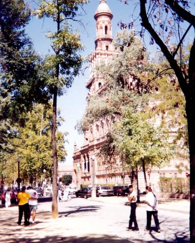 A street scene in Seville showing the Plaza de América with the distinctive Moorish Revival tower of the Museum of Popular Arts and Customs (Museo de Artes y Costumbres Populares) visible behind trees, with pedestrians and cars on the road.