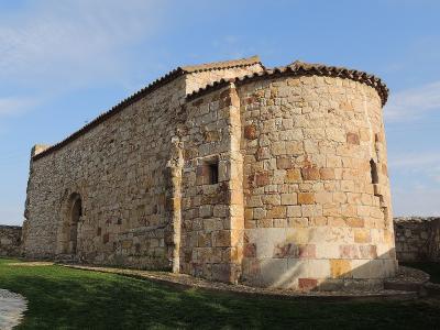 A low-angle exterior shot of a rustic stone church with a rectangular nave and a rounded apse, set against a blue sky with light clouds and surrounded by a low stone wall and grass.