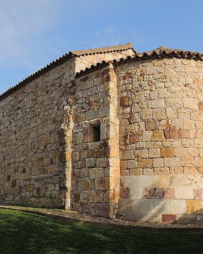 A low-angle exterior shot of a rustic stone church with a rectangular nave and a rounded apse, set against a blue sky with light clouds and surrounded by a low stone wall and grass.