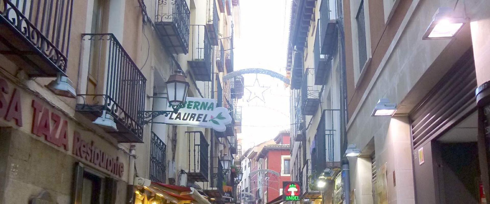 A narrow pedestrian street lined with buildings featuring balconies, outdoor seating for bars, and wooden wine barrels, with a hanging sign reading "CERBERNA LAUREL" and another sign for "LA TAZA".