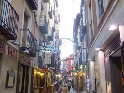 A narrow pedestrian street lined with buildings featuring balconies, outdoor seating for bars, and wooden wine barrels, with a hanging sign reading "CERBERNA LAUREL" and another sign for "LA TAZA".