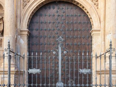 A close-up of an ornate stone church entrance featuring a large arched wooden door with metal studs, flanked by columns and topped with elaborate Baroque-style carvings, viewed through a black wrought-iron gate.