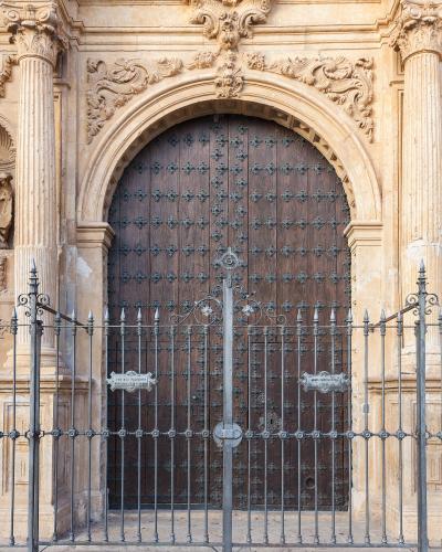 A close-up of an ornate stone church entrance featuring a large arched wooden door with metal studs, flanked by columns and topped with elaborate Baroque-style carvings, viewed through a black wrought-iron gate.
