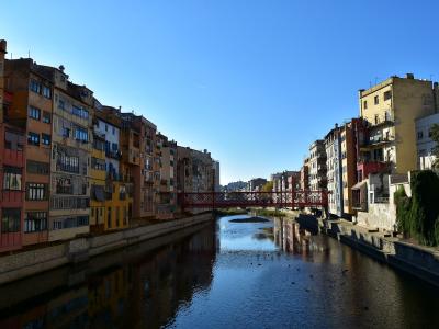 A view of colorful multi-story buildings lining a river with a red metal footbridge crossing it under a clear blue sky.