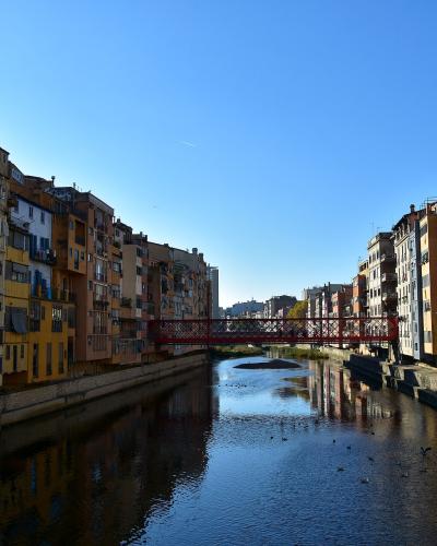 A view of colorful multi-story buildings lining a river with a red metal footbridge crossing it under a clear blue sky.