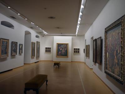 The interior of an art gallery featuring white walls, track lighting, wooden floors, and numerous framed paintings, including a large central altarpiece and a large tapestry or painting on the right wall.