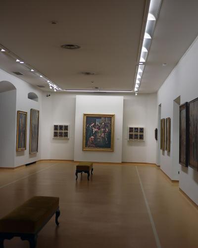 The interior of an art gallery featuring white walls, track lighting, wooden floors, and numerous framed paintings, including a large central altarpiece and a large tapestry or painting on the right wall.
