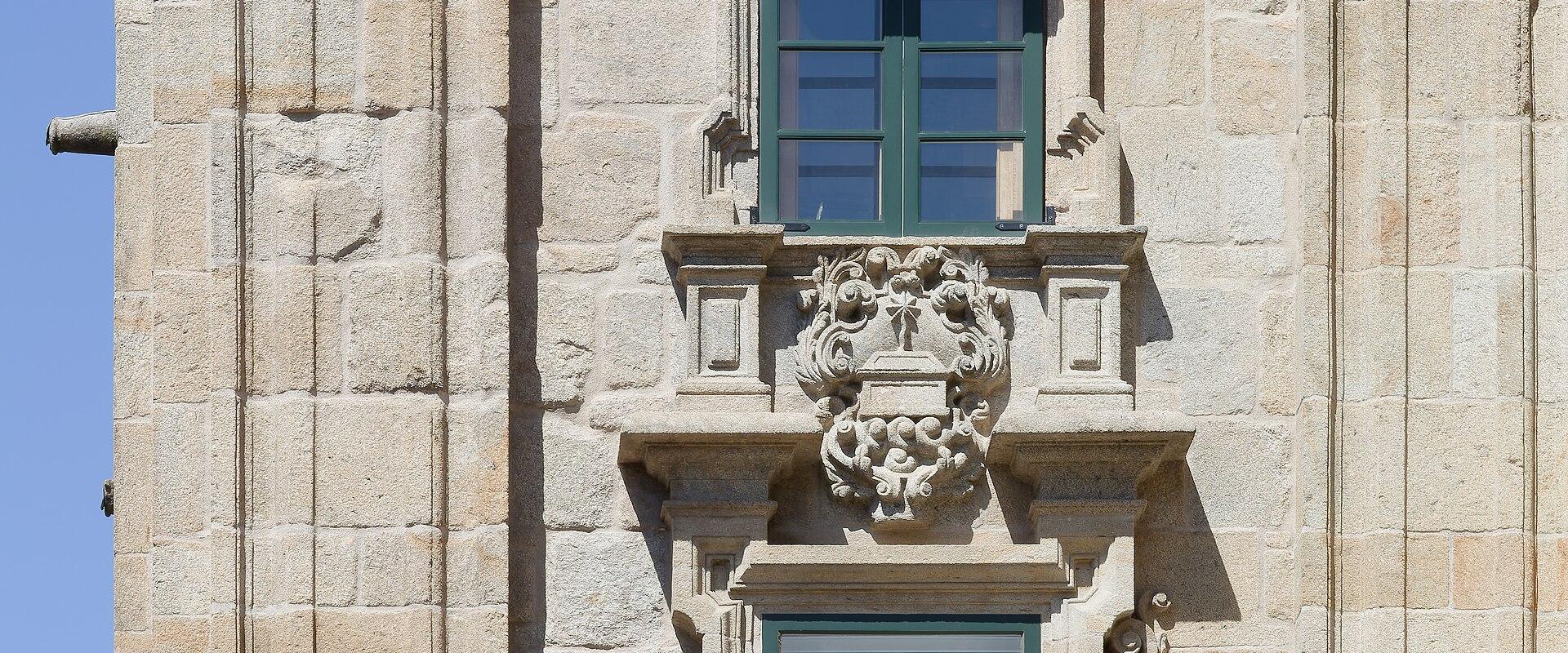 A close-up view of the stone facade of a historic building featuring two vertically aligned windows with green shutters, a wrought-iron balcony, and ornate stone carvings including a coat of arms.
