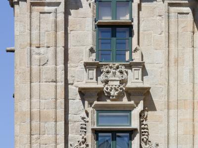 A close-up view of the stone facade of a historic building featuring two vertically aligned windows with green shutters, a wrought-iron balcony, and ornate stone carvings including a coat of arms.