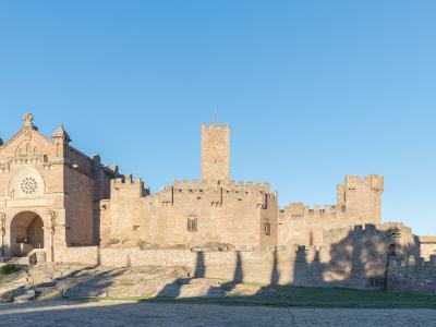 A large, historic stone fortress complex featuring a prominent church facade with a rose window on the left and multiple crenellated towers and walls extending to the right, set against a clear blue sky with hills in the background.