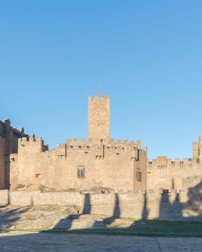 A large, historic stone fortress complex featuring a prominent church facade with a rose window on the left and multiple crenellated towers and walls extending to the right, set against a clear blue sky with hills in the background.