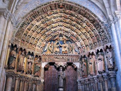 The image shows the interior of a highly ornate Romanesque church portal, featuring a large wooden door, a central tympanum with a sculpted scene of a coronation or enthronement, and multiple concentric archivolts filled with carved figures, flanked 