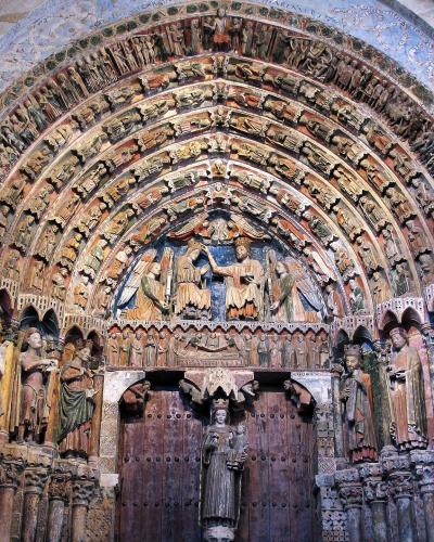 The image shows the interior of a highly ornate Romanesque church portal, featuring a large wooden door, a central tympanum with a sculpted scene of a coronation or enthronement, and multiple concentric archivolts filled with carved figures, flanked 