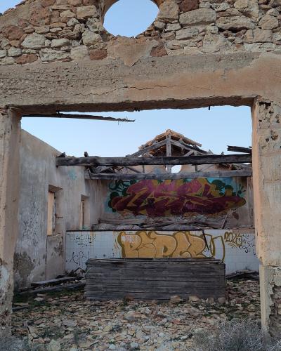 A ruined stone building with a large doorway, graffiti-covered interior walls, and a blue informational plaque mounted on the left wall.