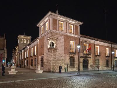 A night photograph of a large, historic brick and stone building with illuminated windows, balconies, and a Spanish flag hanging from the facade, situated on a cobblestone plaza with pedestrians and streetlights.