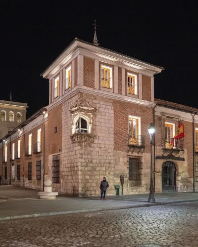 A night photograph of a large, historic brick and stone building with illuminated windows, balconies, and a Spanish flag hanging from the facade, situated on a cobblestone plaza with pedestrians and streetlights.