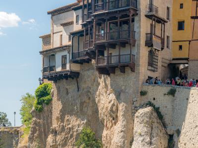 A cluster of historic stone houses with distinctive wooden balconies built directly onto the edge of a steep, rocky cliff face under a blue sky.