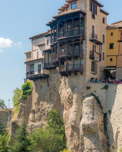 A cluster of historic stone houses with distinctive wooden balconies built directly onto the edge of a steep, rocky cliff face under a blue sky.