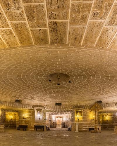 The image shows the interior of a stone crypt or underground chapel with a large central dome, arched ceilings, religious paintings, and candle sconces.