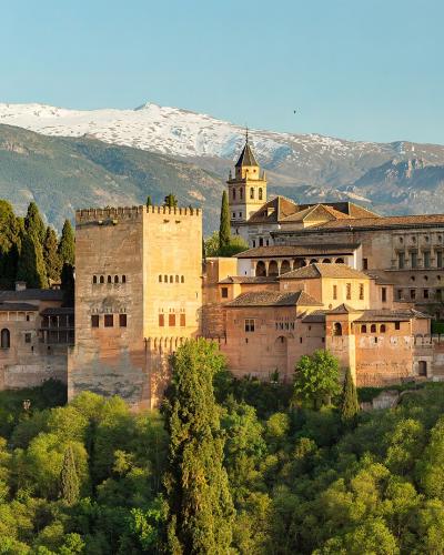 A panoramic view of the Alhambra palace complex, featuring the Palace of Charles V and the Alcazaba fortress, set against the snow-capped Sierra Nevada mountains.