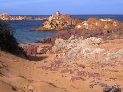A coastal scene showing a sandy beach with reddish-brown and white rock formations, turquoise water, and a prominent jagged rock outcrop in the sea under a clear blue sky.