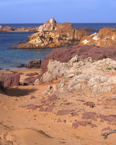 A coastal scene showing a sandy beach with reddish-brown and white rock formations, turquoise water, and a prominent jagged rock outcrop in the sea under a clear blue sky.