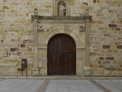 A close-up view of a stone church facade featuring a large arched wooden door, flanked by columns with sculpted figures, and topped by a niche containing a statue of a seated saint.