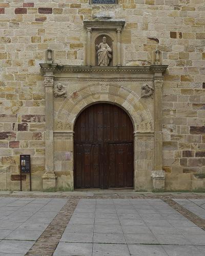 A close-up view of a stone church facade featuring a large arched wooden door, flanked by columns with sculpted figures, and topped by a niche containing a statue of a seated saint.