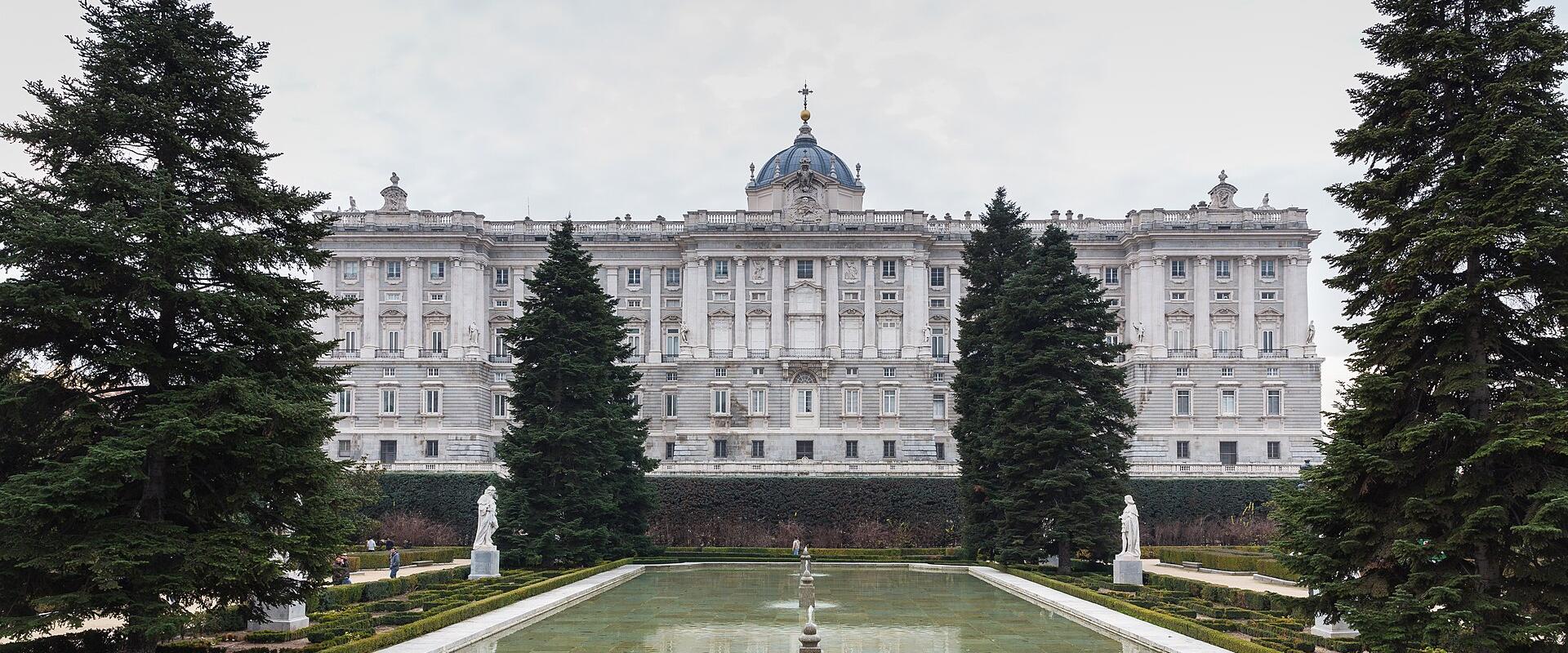 A wide shot of a massive, ornate white palace facade with a central blue dome and cross, viewed from a formal garden featuring a long rectangular reflecting pool, flanked by tall evergreen trees and statues.