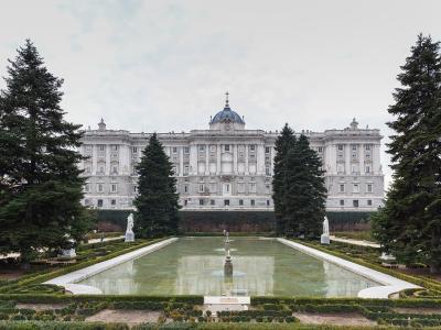 A wide shot of a massive, ornate white palace facade with a central blue dome and cross, viewed from a formal garden featuring a long rectangular reflecting pool, flanked by tall evergreen trees and statues.