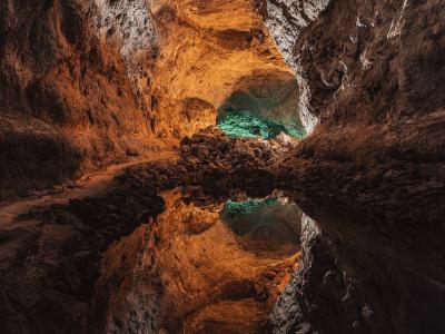 A high-angle view of a paved stone pathway leading to the dark, rough-hewn entrance of a volcanic cave, surrounded by arid volcanic rock and sparse vegetation.