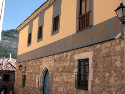 A two-story building with a stone lower facade and yellow upper walls, featuring a blue arched door, barred windows, and a long horizontal inscription in large orange letters on a grey band; two people stand on the street to the left.