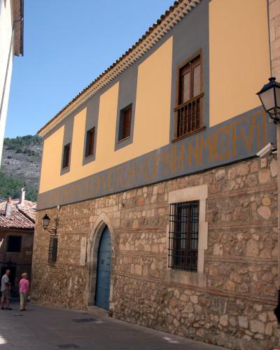 A two-story building with a stone lower facade and yellow upper walls, featuring a blue arched door, barred windows, and a long horizontal inscription in large orange letters on a grey band; two people stand on the street to the left.