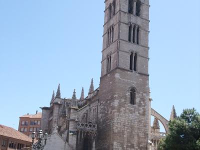 A low-angle photograph of a large stone church with a tall, square bell tower featuring arched openings and a pyramidal tiled roof, set against a clear blue sky with a street lamp and bicycles in the foreground.