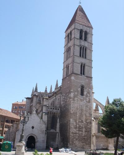 A low-angle photograph of a large stone church with a tall, square bell tower featuring arched openings and a pyramidal tiled roof, set against a clear blue sky with a street lamp and bicycles in the foreground.