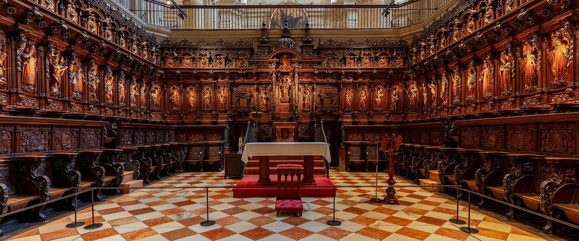 The image shows the interior of a cathedral featuring an elaborate, dark wood choir stall (retablo) surrounding a central altar on a red carpet, set against a floor with a distinctive red and white diamond-patterned tile design.