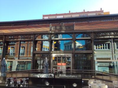 The image shows the exterior of a large, historic iron-and-glass market building with a terracotta-tiled roof, featuring outdoor seating areas and a prominent sign reading "MERCADO DEL VAL" above the entrance.