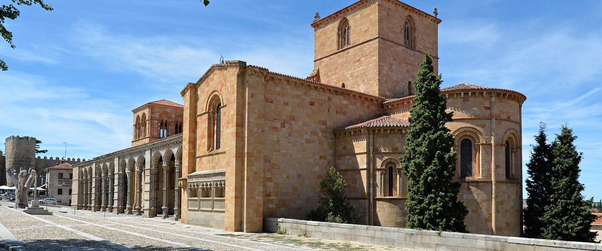A large Romanesque stone basilica with a prominent tower, apse, and long arcade of arches on the left, set against a blue sky with a stone wall and tower visible in the background.