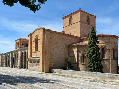 A large Romanesque stone basilica with a prominent tower, apse, and long arcade of arches on the left, set against a blue sky with a stone wall and tower visible in the background.