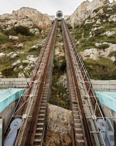 A view looking up a steep, dual-track funicular railway ascending a rocky, vegetated hillside, with a spherical glass cabin visible at the top station.