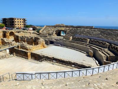 A large, semi-circular Roman amphitheatre ruin with tiered stone seating, a sandy arena floor, and modern metal railings, set against a blue sky and sea with a multi-story building in the background.