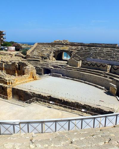 A large, semi-circular Roman amphitheatre ruin with tiered stone seating, a sandy arena floor, and modern metal railings, set against a blue sky and sea with a multi-story building in the background.