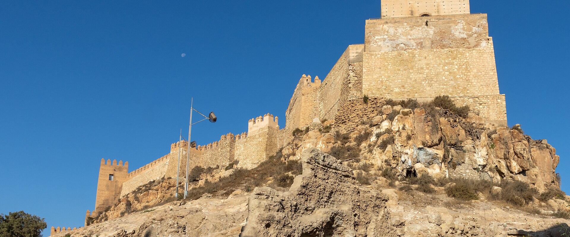 A low-angle view of a large, fortified Moorish citadel with crenellated walls and a prominent rectangular tower (Torre de la Vela) situated on a rocky hill under a clear blue sky.