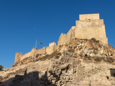 A low-angle view of a large, fortified Moorish citadel with crenellated walls and a prominent rectangular tower (Torre de la Vela) situated on a rocky hill under a clear blue sky.